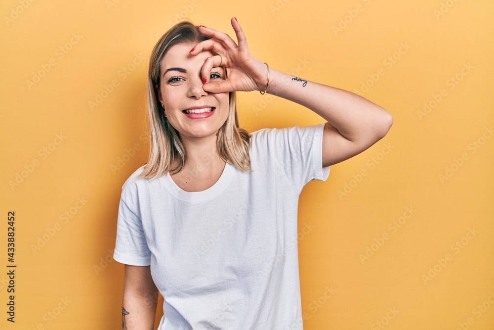 Beautiful caucasian woman wearing casual white t shirt doing ok gesture with hand smiling, eye looking through fingers with happy face. Beautiful caucasian woman wearing casual white t shirt doing ok gesture with hand smiling, eye looking through fingers with happy face.