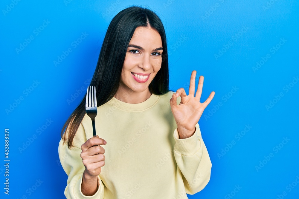 Young brunette woman holding one silver fork doing ok sign with fingers, smiling friendly gesturing excellent symbol Young brunette woman holding one silver fork doing ok sign with fingers, smiling friendly gesturing excellent symbol