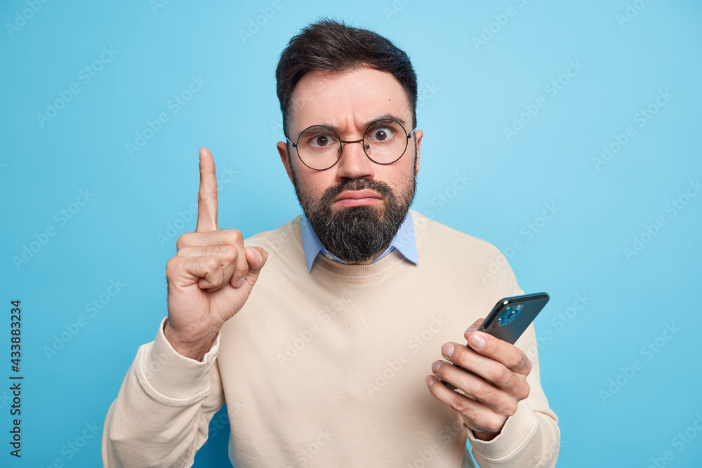 Strict serious bearded adult man raises index finger has excellent idea uses new mobile application holds smartphone wears spectacles and sweater isolated over blue backgound. Technology user Strict serious bearded adult man raises index finger has excellent idea uses new mobile application holds smartphone wears spectacles and sweater isolated over blue backgound. Technology user