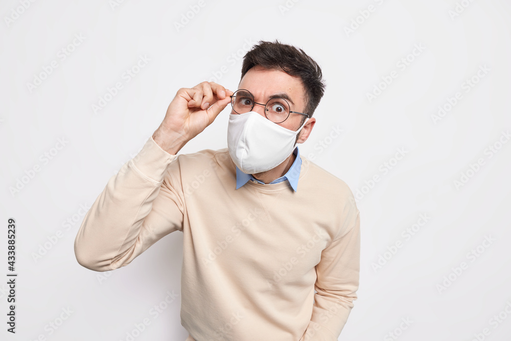 Serious attentive brunet man wears protective mask in public place keeps hand on spectacles prevents himself from coronavirus dressed casually isolated over white background. Covid 19 outbreak Serious attentive brunet man wears protective mask in public place keeps hand on spectacles prevents himself from coronavirus dressed casually isolated over white background. Covid 19 outbreak
