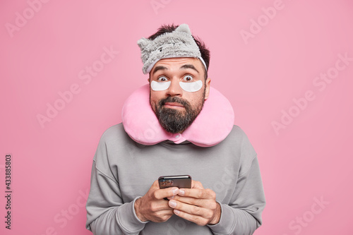 Confused surprised man with beard uses neck pillow for better sleep applies beauty patches under eyes holds smartphone poses indoor against pink background avoids stiffness going to have nap © Wayhome Studio