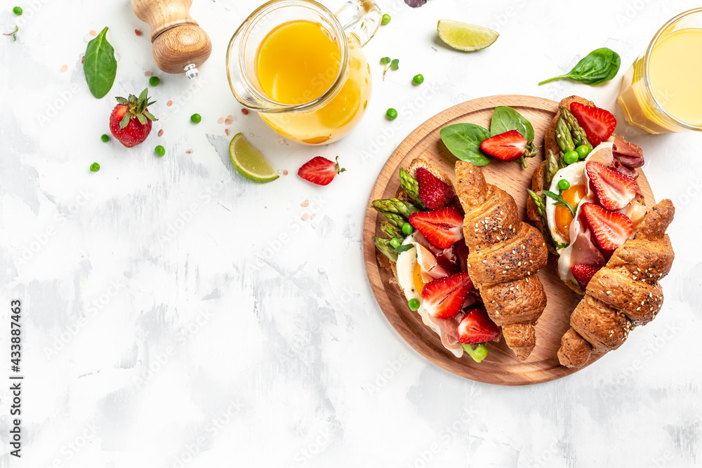 traditional breakfast fresh croissants with avocado, asparagus, jamon, ham, prosciutto, strawberries, sunny side up eggs and juice. banner format, top view