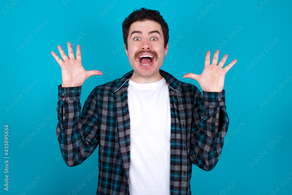 Naklejka premium young Caucasian man with moustache wearing plaid shirt against blue wall raising hands up, having eyes full of happiness rejoicing his great achievements. Achievement, success concept.