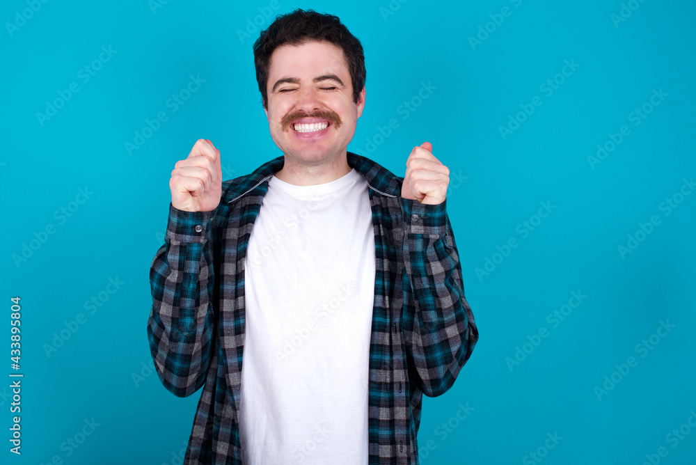 young Caucasian man with moustache wearing plaid shirt against blue wall rejoicing his success and victory clenching fists with joy being happy to achieve aim and goals. Positive emotions, feelings. young Caucasian man with moustache wearing plaid shirt against blue wall rejoicing his success and victory clenching fists with joy being happy to achieve aim and goals. Positive emotions, feelings.
