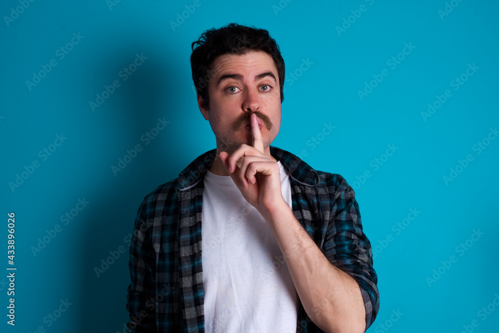 young Caucasian man with moustache wearing plaid shirt against blue wall makes hush gesture, asks be quiet. Don't tell my secret or not speak too loud, please! young Caucasian man with moustache wearing plaid shirt against blue wall makes hush gesture, asks be quiet. Don't tell my secret or not speak too loud, please!