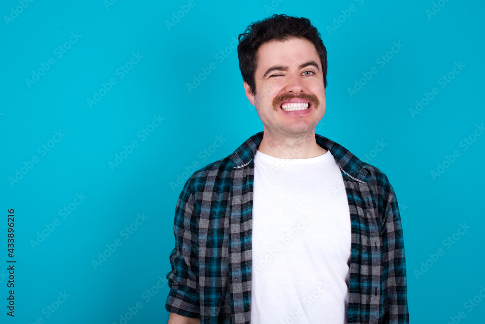young Caucasian man with moustache wearing plaid shirt against blue wall blinking eyes with pleasure having happy expression. Facial expressions and people emotions concept. young Caucasian man with moustache wearing plaid shirt against blue wall blinking eyes with pleasure having happy expression. Facial expressions and people emotions concept.