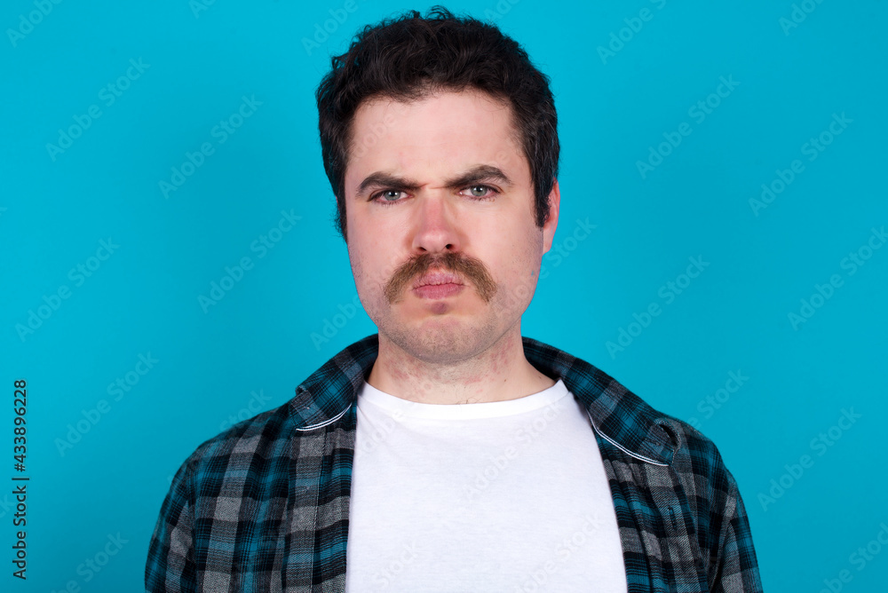 Fototapeta premium young Caucasian man with moustache wearing plaid shirt against blue wall frowning his eyebrows being displeased with something.
