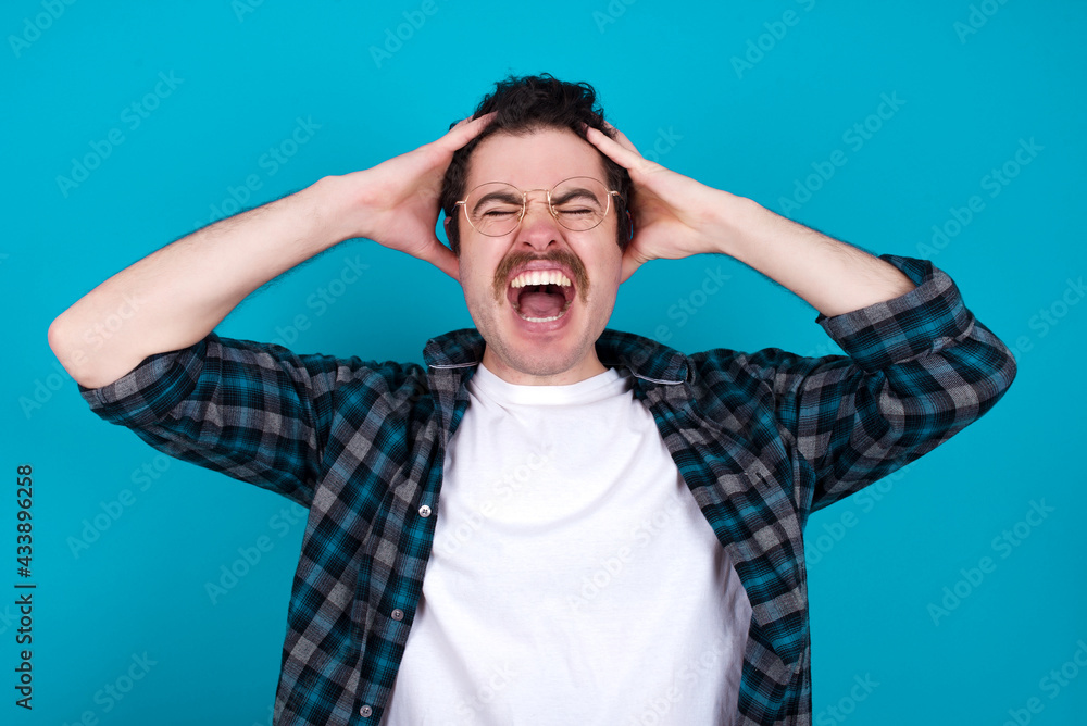 Shocked panic young Caucasian man with moustache wearing plaid shirt against blue wall holding hands on head and screaming in despair and frustration. Shocked panic young Caucasian man with moustache wearing plaid shirt against blue wall holding hands on head and screaming in despair and frustration.