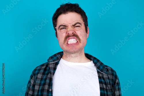 Mad crazy young Caucasian man with moustache wearing plaid shirt against blue wall clenches teeth angrily, being annoyed with coming noise. Negative feeling concept.