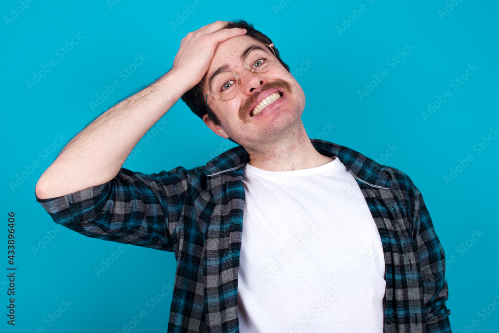 Oops, what did I do? young Caucasian man with moustache wearing plaid shirt against blue wall holding hand on forehead with frightened and regret expression. Oops, what did I do? young Caucasian man with moustache wearing plaid shirt against blue wall holding hand on forehead with frightened and regret expression.