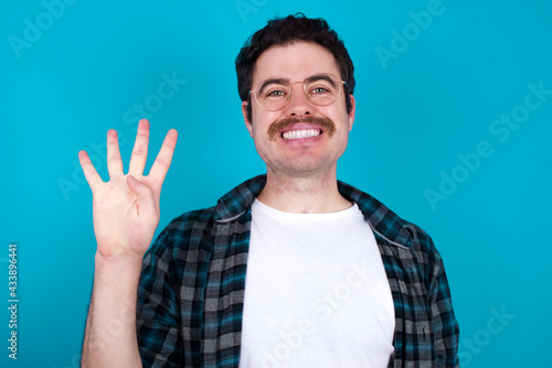 young Caucasian man with moustache wearing plaid shirt against blue wall showing and pointing up with fingers number four while smiling confident and happy.