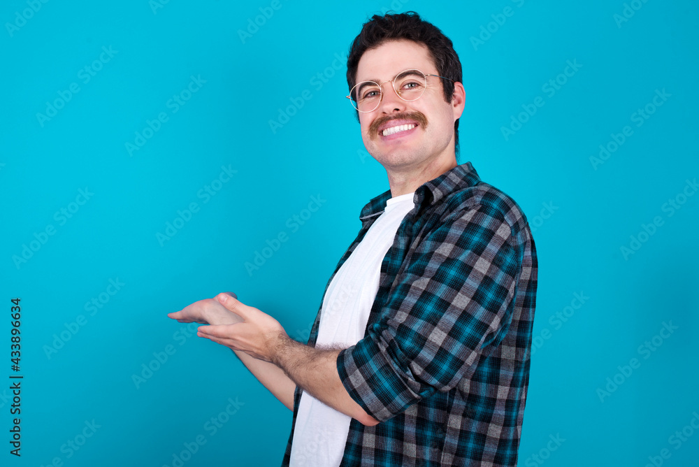 young Caucasian man with moustache wearing plaid shirt against blue wall Inviting to enter smiling natural with open hands. Welcome sign. young Caucasian man with moustache wearing plaid shirt against blue wall Inviting to enter smiling natural with open hands. Welcome sign.