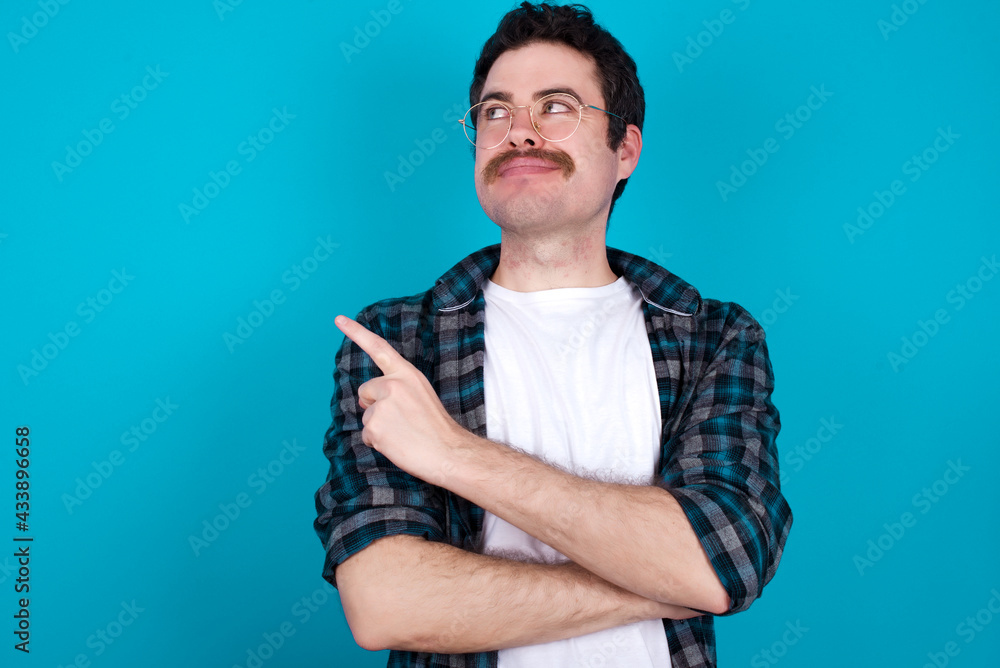 Portrait of young Caucasian man with moustache wearing plaid shirt against blue wall posing on camera with tricky look, presenting product with index finger. Advertisement concept. Portrait of young Caucasian man with moustache wearing plaid shirt against blue wall posing on camera with tricky look, presenting product with index finger. Advertisement concept.