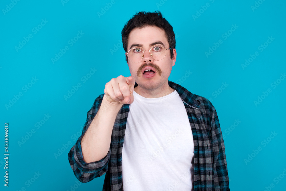 Shocked young Caucasian man with moustache wearing plaid shirt against blue wall points front with index finger at camera and. Surprise and advertisement concept. Shocked young Caucasian man with moustache wearing plaid shirt against blue wall points front with index finger at camera and. Surprise and advertisement concept.