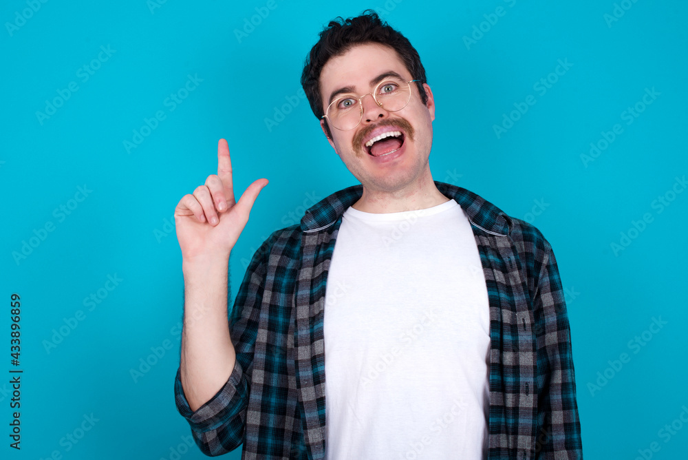 Pleasant looking young Caucasian man with moustache wearing plaid shirt against blue wall has clever expression, raises one finger, remembers himself not to forget tell important thing. Pleasant looking young Caucasian man with moustache wearing plaid shirt against blue wall has clever expression, raises one finger, remembers himself not to forget tell important thing.