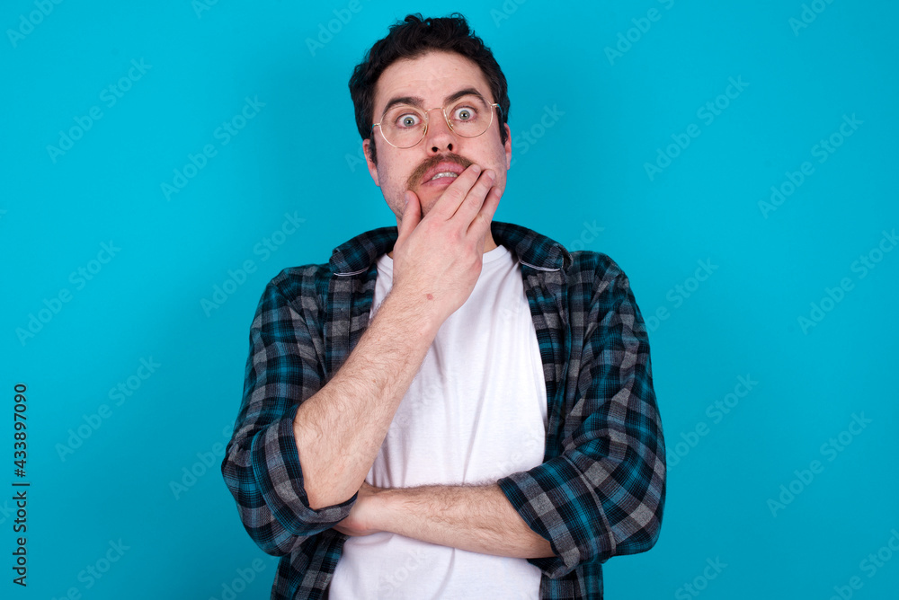 young Caucasian man with moustache wearing plaid shirt against blue wall covering mouth with hands scared from something or someone bitting nails young Caucasian man with moustache wearing plaid shirt against blue wall covering mouth with hands scared from something or someone bitting nails