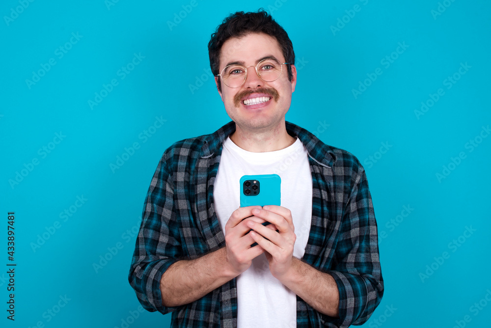 Smiling young Caucasian man with moustache wearing plaid shirt against blue wall friendly and happily holding mobile phone taking selfie in mirror. Smiling young Caucasian man with moustache wearing plaid shirt against blue wall friendly and happily holding mobile phone taking selfie in mirror.