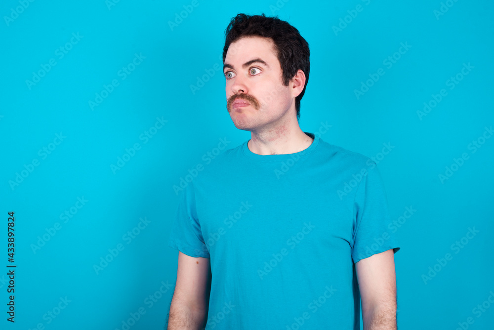 young handsome Caucasian man with moustache wearing blue t-shirt against blue background stares aside with wondered expression has speechless expression. Embarrassed model looks in surprise young handsome Caucasian man with moustache wearing blue t-shirt against blue background stares aside with wondered expression has speechless expression. Embarrassed model looks in surprise