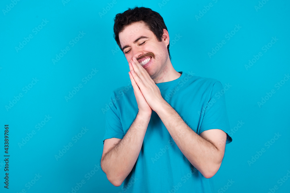 Overjoyed young handsome Caucasian man with moustache wearing blue t-shirt against blue background laughs joyfully and keeps palms pressed together hears something funny Overjoyed young handsome Caucasian man with moustache wearing blue t-shirt against blue background laughs joyfully and keeps palms pressed together hears something funny