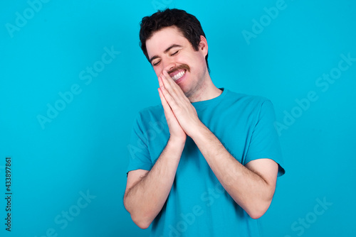 Overjoyed young handsome Caucasian man with moustache wearing blue t-shirt against blue background laughs joyfully and keeps palms pressed together hears something funny
