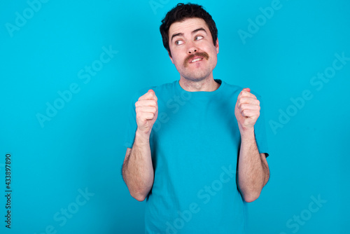 young handsome Caucasian man with moustache wearing blue t-shirt against blue background clenches fists and awaits for something nice happened looks away bites lips and waits announcement of results