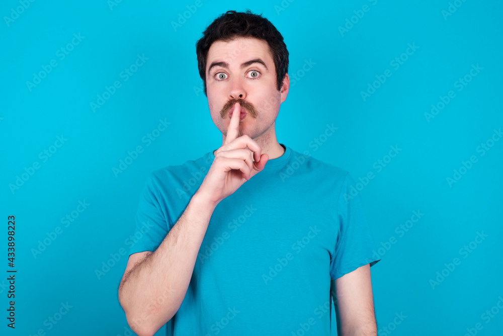 Surprised young handsome Caucasian man with moustache wearing blue t-shirt against blue background makes silence gesture, keeps finger over lips and looks mysterious at camera Surprised young handsome Caucasian man with moustache wearing blue t-shirt against blue background makes silence gesture, keeps finger over lips and looks mysterious at camera