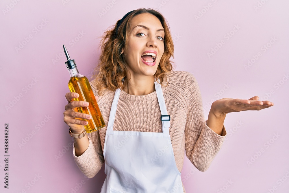 Young caucasian woman wearing apron holding olive oil can celebrating achievement with happy smile and winner expression with raised hand Young caucasian woman wearing apron holding olive oil can celebrating achievement with happy smile and winner expression with raised hand