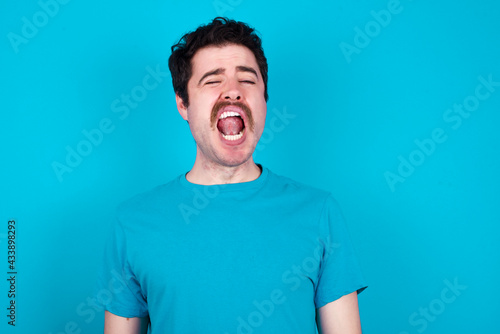 young handsome Caucasian man with moustache wearing blue t-shirt against blue background yawns with opened mouth stands. Daily morning routine