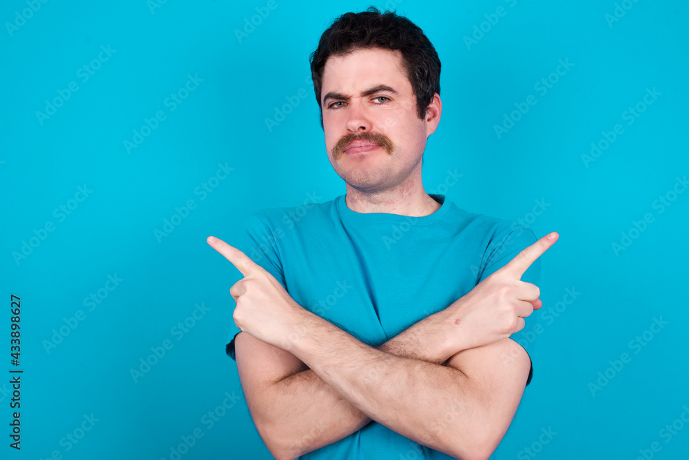 Serious young handsome Caucasian man with moustache wearing blue t-shirt against blue background crosses hands and points at different sides hesitates between two items. Hard decision concept Serious young handsome Caucasian man with moustache wearing blue t-shirt against blue background crosses hands and points at different sides hesitates between two items. Hard decision concept