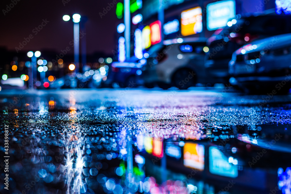 Rainy night in the parking shopping mall, rows of parked cars in the light of advertising.  Close up view of a puddle on the asphalt level