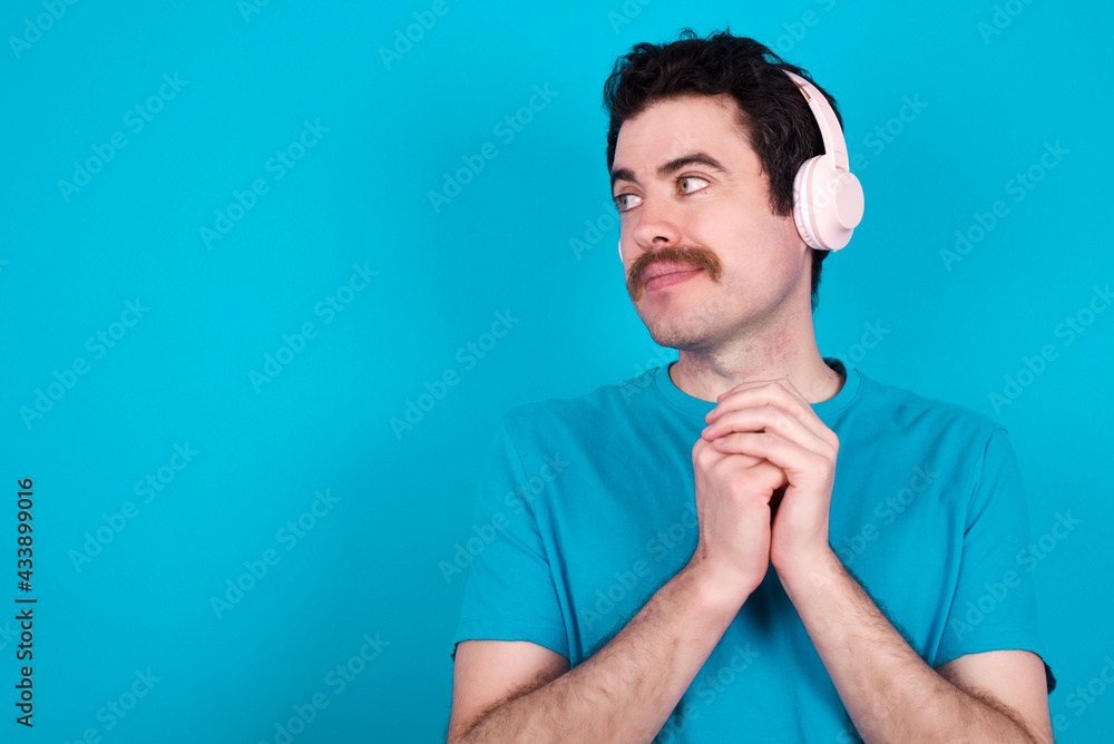 young handsome Caucasian man with moustache wearing blue t-shirt against blue background wears stereo headphones listening to music concentrated and looking aside with interest. young handsome Caucasian man with moustache wearing blue t-shirt against blue background wears stereo headphones listening to music concentrated and looking aside with interest.