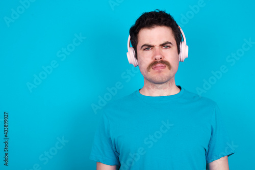 Wallpaper Mural Serious displeased young handsome Caucasian man with moustache wearing blue t-shirt against blue wall looks puzzled at camera being angry wears stereo headphones listens music while walking at street Torontodigital.ca