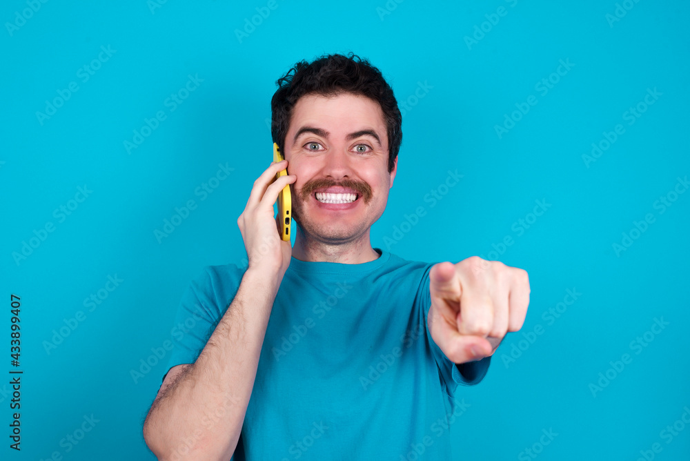 Positive young handsome Caucasian man with moustache wearing blue t-shirt against blue indicates directly at camera has telephone conversation smiles broadly enjoys talking long hours. You join me Positive young handsome Caucasian man with moustache wearing blue t-shirt against blue indicates directly at camera has telephone conversation smiles broadly enjoys talking long hours. You join me