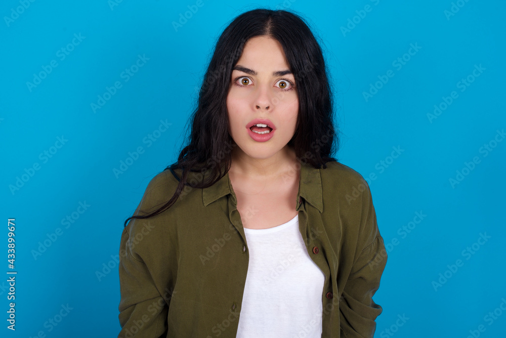 Obraz premium young beautiful tattooed girl standing against blue background having stunned and shocked look, with mouth open and jaw dropped exclaiming: Wow, I can't believe this. Surprise and shock