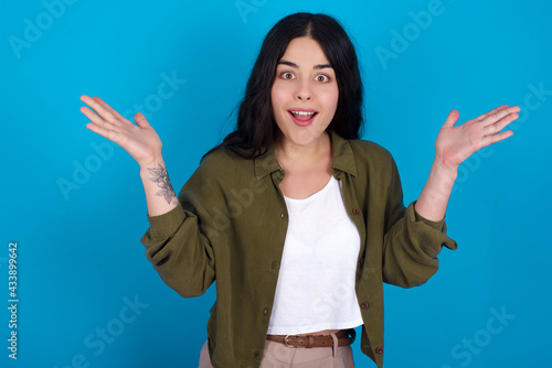 young beautiful tattooed girl standing against blue background raising hands up, having eyes full of happiness rejoicing his great achievements. Achievement, success concept.