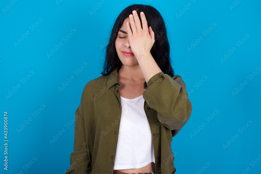 Frustrated young beautiful tattooed girl standing against blue background holding hand on forehead being depressed regretting what he did having headache, looking stressful. Frustrated young beautiful tattooed girl standing against blue background holding hand on forehead being depressed regretting what he did having headache, looking stressful.
