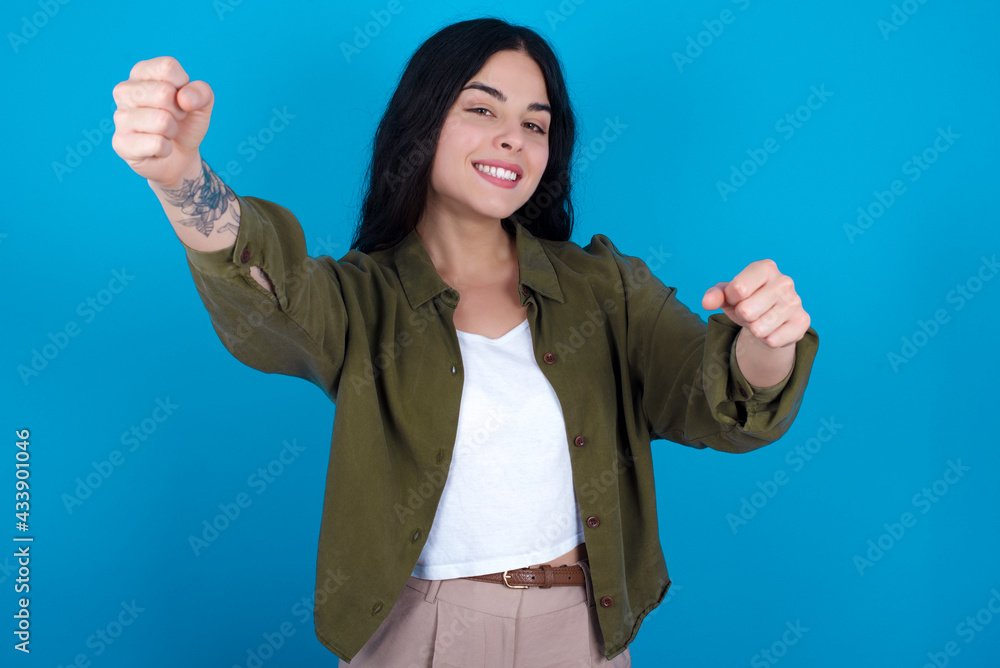 Portrait of charming young beautiful tattooed girl standing against blue background, smiling broadly while holding hands over her head. Confidence and relax concept. Portrait of charming young beautiful tattooed girl standing against blue background, smiling broadly while holding hands over her head. Confidence and relax concept.