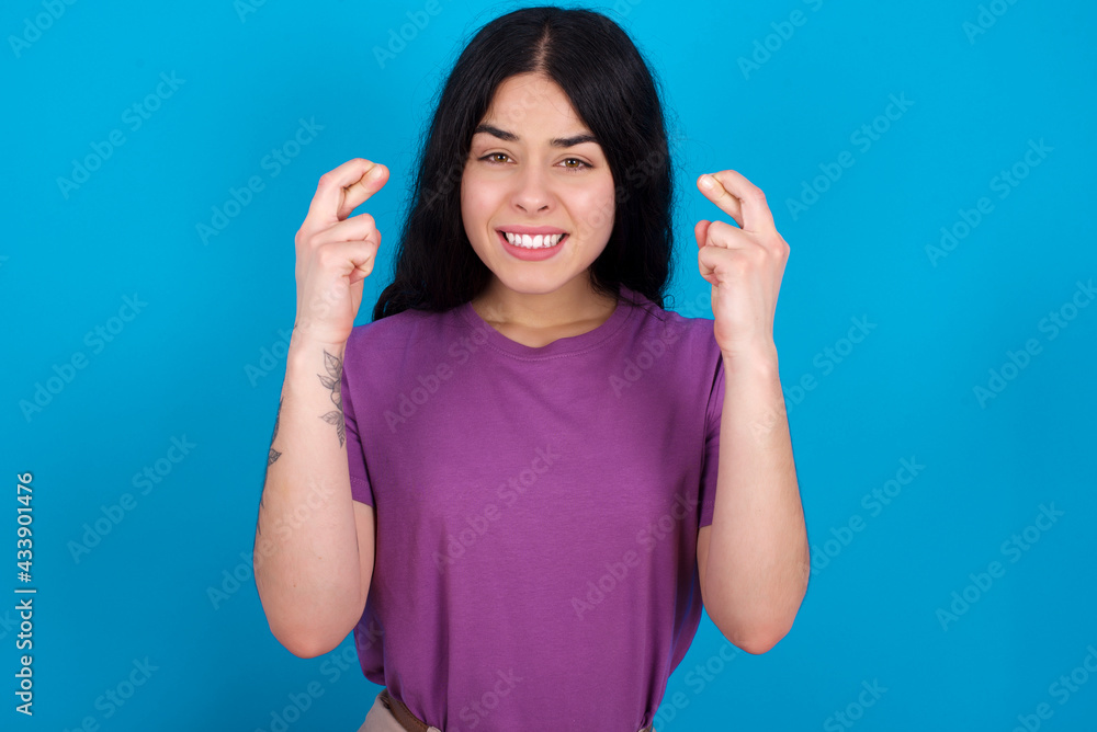 young beautiful tattooed girl wearing purple t-shirt standing against blue background holding fingers crossed with worried expression hoping boss didn't noticed mistakes at work. young beautiful tattooed girl wearing purple t-shirt standing against blue background holding fingers crossed with worried expression hoping boss didn't noticed mistakes at work.