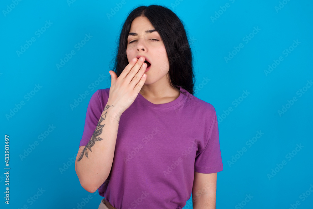 Sleepy young beautiful tattooed girl wearing purple t-shirt standing against blue background yawning with messy hair, feeling tired after sleepless night, yawning, covering mouth with palm. Sleepy young beautiful tattooed girl wearing purple t-shirt standing against blue background yawning with messy hair, feeling tired after sleepless night, yawning, covering mouth with palm.