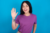 young beautiful tattooed girl wearing purple t-shirt standing against blue background waiving saying hello or goodbye happy and smiling, friendly welcome gesture.
