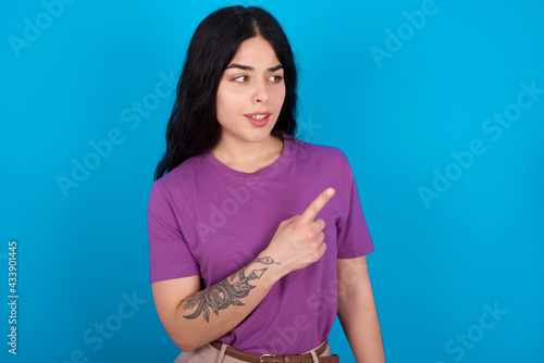 Happy cheerful smiling young beautiful tattooed girl wearing purple t-shirt standing against blue background looking and pointing aside with hand. Copy space and advertisement concept.