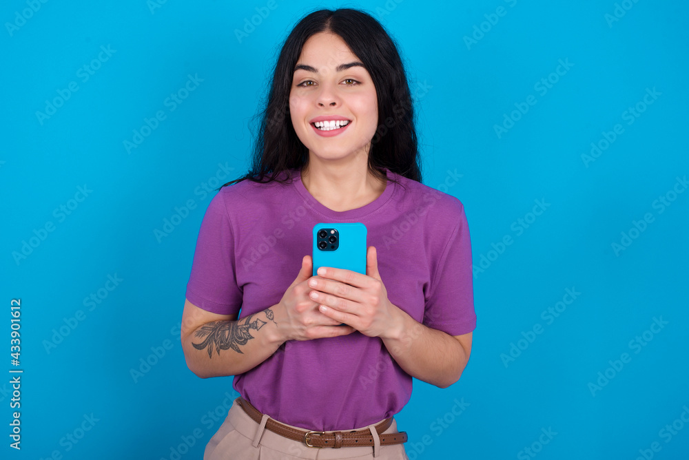 Smiling young beautiful tattooed girl wearing purple t-shirt standing against blue background friendly and happily holding mobile phone taking selfie in mirror. Smiling young beautiful tattooed girl wearing purple t-shirt standing against blue background friendly and happily holding mobile phone taking selfie in mirror.