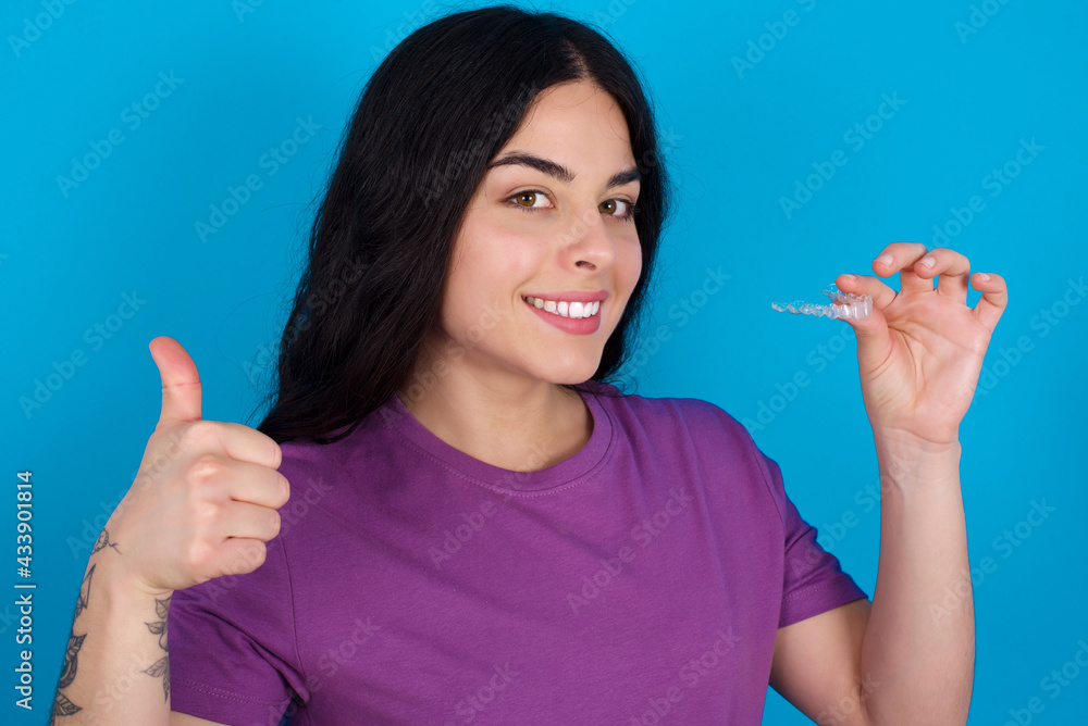young beautiful tattooed girl wearing purple t-shirt standing against blue wall holding an invisible braces aligner and rising thumb up, recommending this new treatment. Dental healthcare concept. young beautiful tattooed girl wearing purple t-shirt standing against blue wall holding an invisible braces aligner and rising thumb up, recommending this new treatment. Dental healthcare concept.