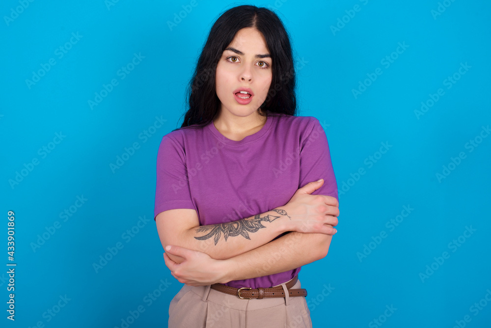 Shocked embarrassed young beautiful tattooed girl wearing purple t-shirt standing against blue background keeps mouth widely opened. Hears unbelievable novelty stares in stupor Shocked embarrassed young beautiful tattooed girl wearing purple t-shirt standing against blue background keeps mouth widely opened. Hears unbelievable novelty stares in stupor