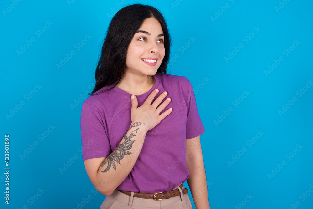 Joyful young beautiful tattooed girl wearing purple t-shirt standing against blue background expresses positive emotions recalls something funny keeps hand on chest and giggles happily.