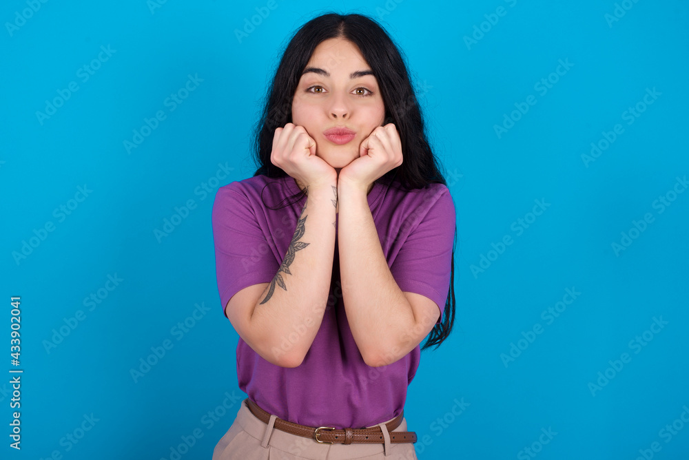 young beautiful tattooed girl wearing purple t-shirt standing against blue background with surprised expression keeps hands under chin keeps lips folded makes funny grimace young beautiful tattooed girl wearing purple t-shirt standing against blue background with surprised expression keeps hands under chin keeps lips folded makes funny grimace