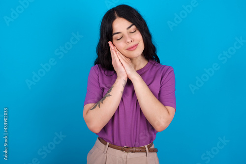 young beautiful tattooed girl wearing purple t-shirt standing against blue background leans on pressed palms closes eyes and has pleasant smile dreams about something © Jihan