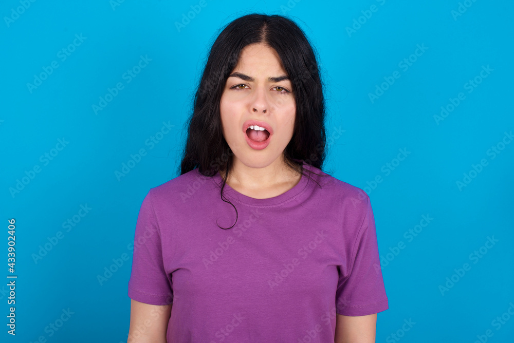 Fototapeta premium young beautiful tattooed girl wearing blue t-shirt standing against blue background yawns with opened mouth stands. Daily morning routine