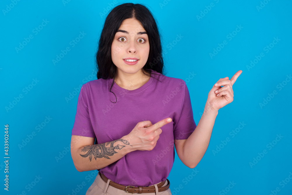 young beautiful tattooed girl wearing blue t-shirt standing against blue background points at copy space indicates for advertising gives right direction young beautiful tattooed girl wearing blue t-shirt standing against blue background points at copy space indicates for advertising gives right direction