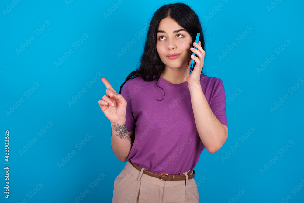 young beautiful tattooed girl wearing blue t-shirt standing against blue background speaks on mobile phone spends free time indoors calls to friend. young beautiful tattooed girl wearing blue t-shirt standing against blue background speaks on mobile phone spends free time indoors calls to friend.